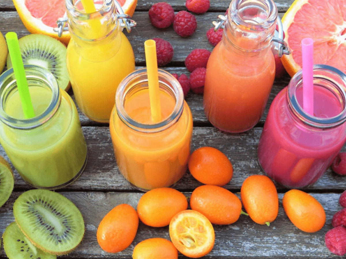 Green, orange, red and yellow glasses of juice with a variety of fruits arranged around them.