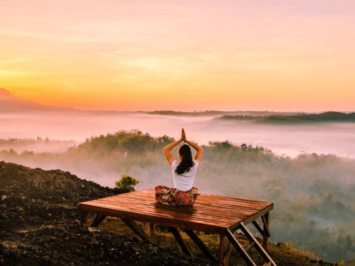 A woman sitting on a wooden bench in a yoga position overlooking a misty forest from above