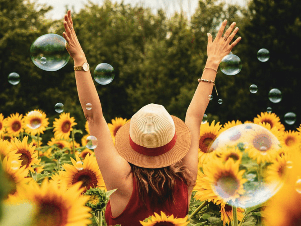 A woman with her hands in the air, wearing a large-brimmed hat standing in a field of tall sunflowers