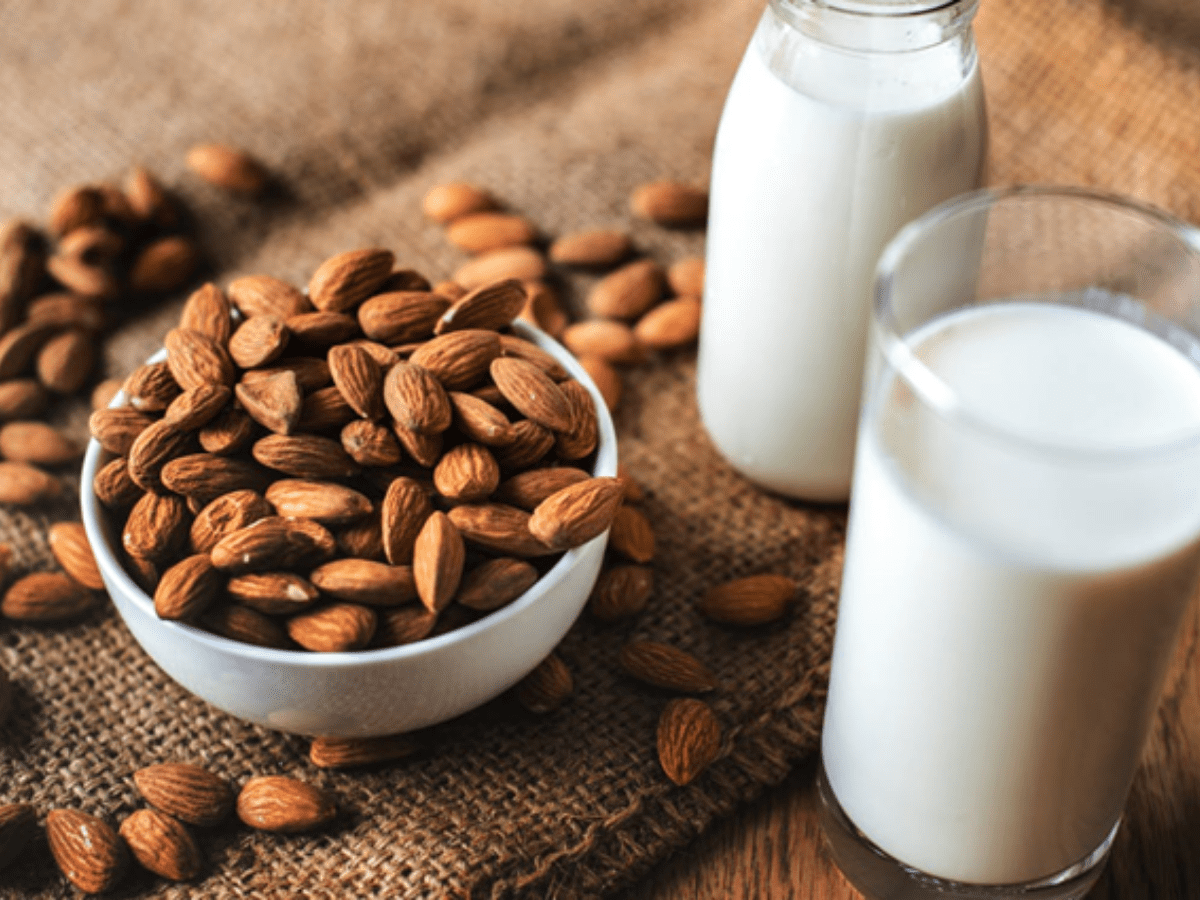 Bowl of almonds with almonds scattered around the sides and a bottle and glass of milk