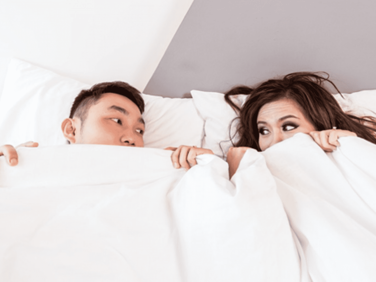 Man in woman in bed with white covers pulled up to their noses, looking at each other.