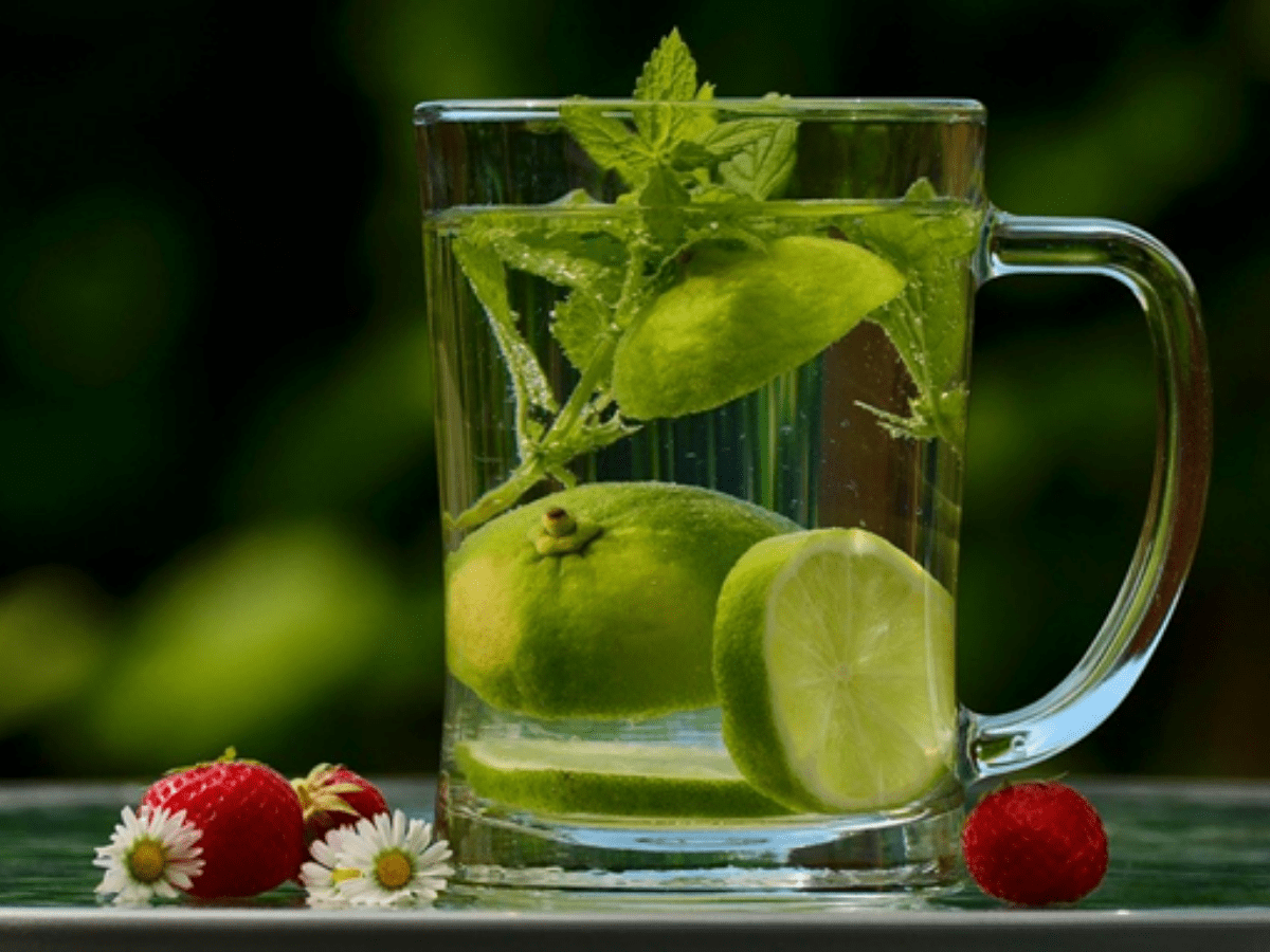 Clear cup of water with slices of lime and lemon balm in it. Raspberries on the table next to cup.